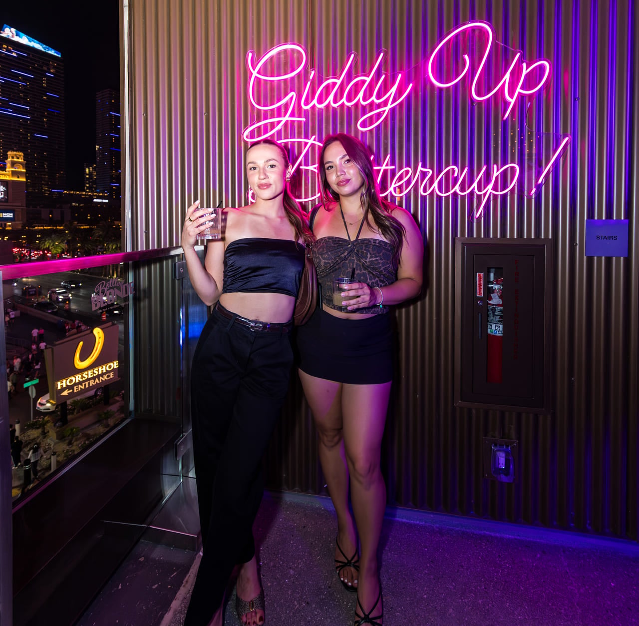 Two women pose for a photo on a rooftop at night next to a neon sign that reads "Giddy Up Buttercup!"