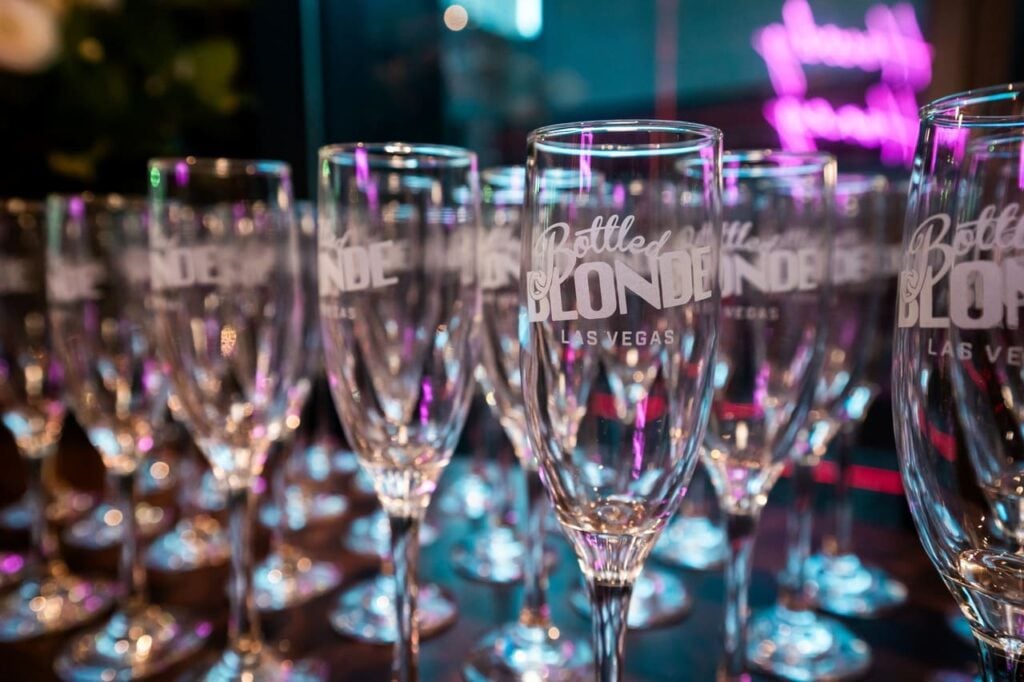Close-up of champagne flutes etched with the Bottled Blonde Las Vegas logo, arranged in rows with colorful lights reflecting on the glass.