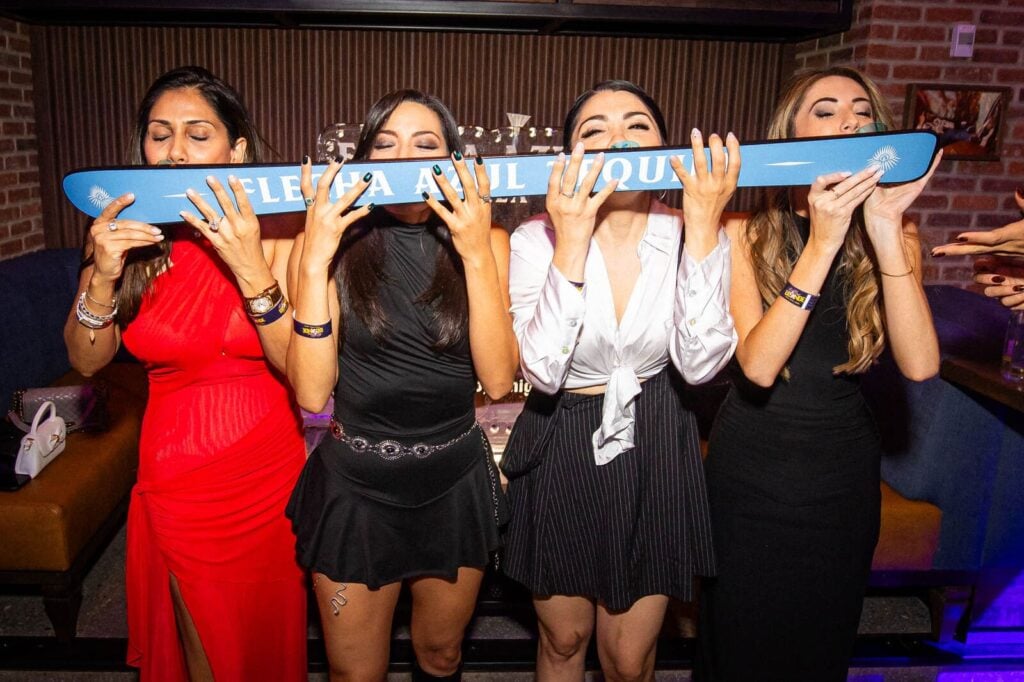 Four women in party attire taking a communal shot from a branded ski at a nightlife venue