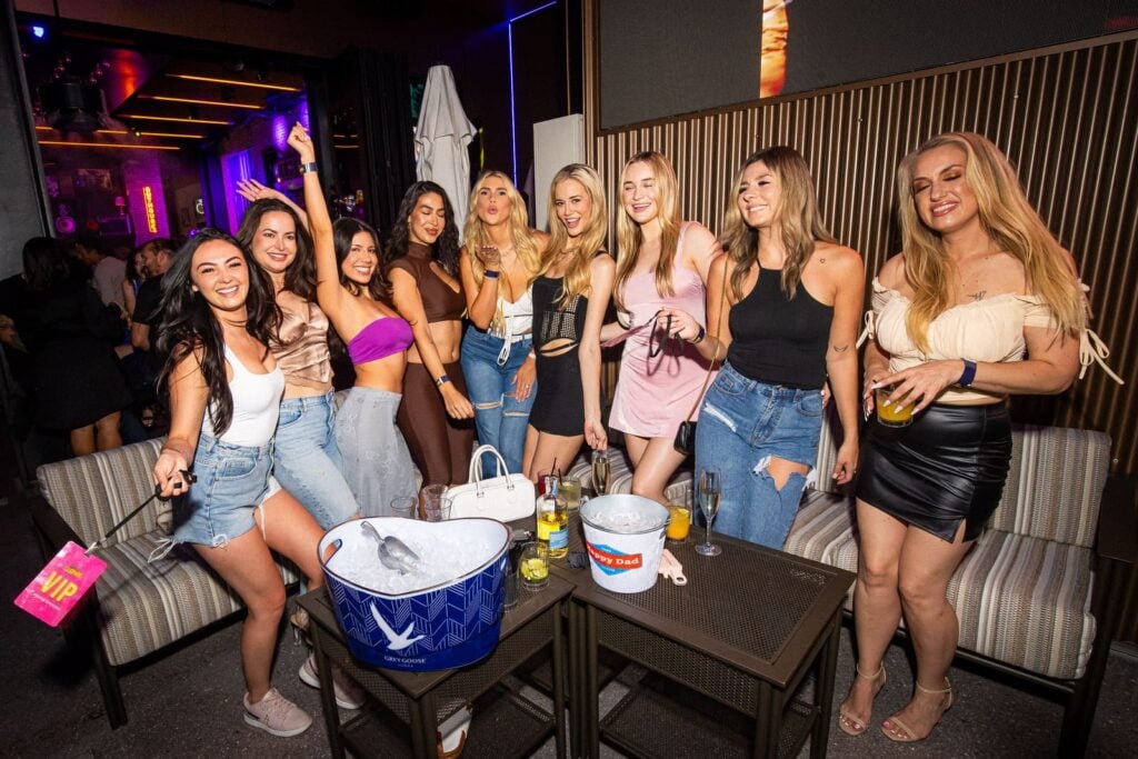 Group of women in casual nightlife attire smiling and posing on a rooftop patio with drink buckets