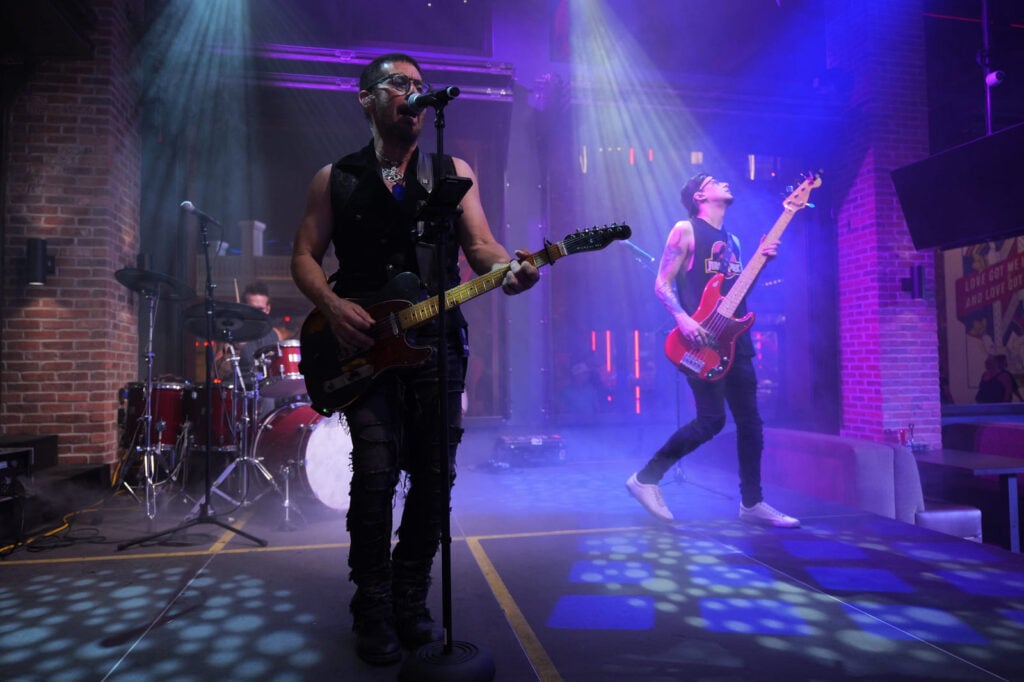 Guitarist singing with bassist on stage under purple lights