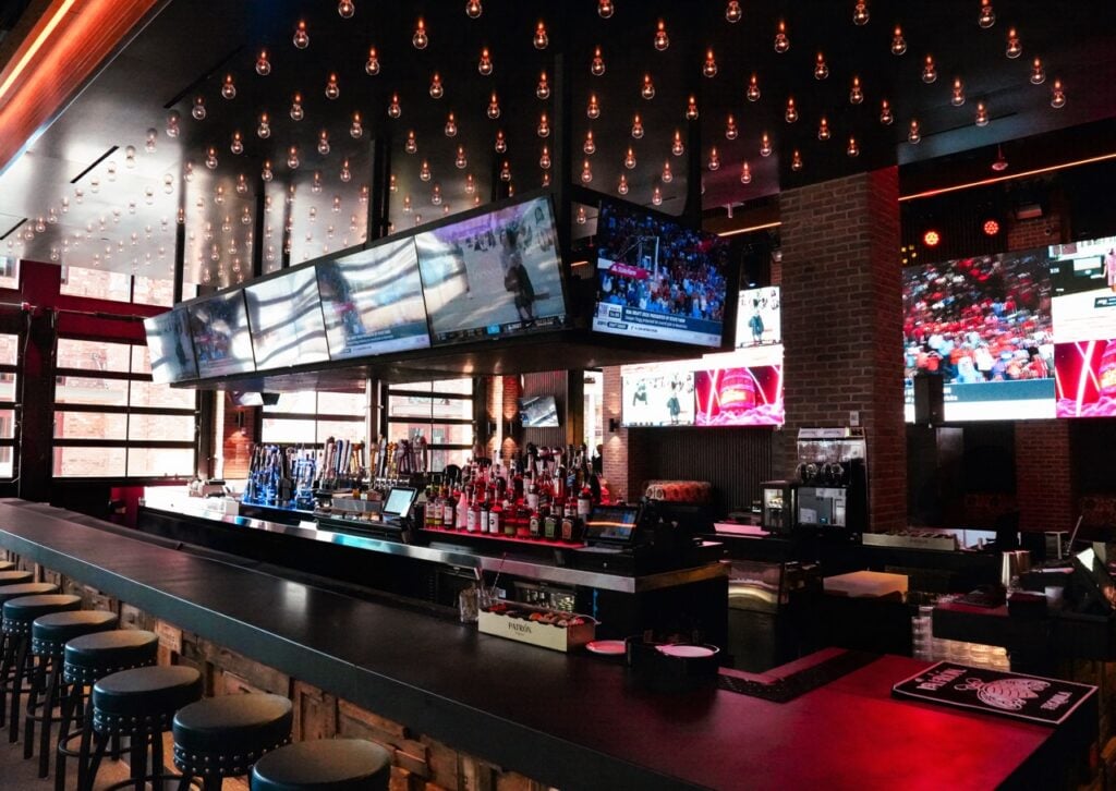 Long bar counter with liquor bottles multiple TV screens and hanging light bulbs