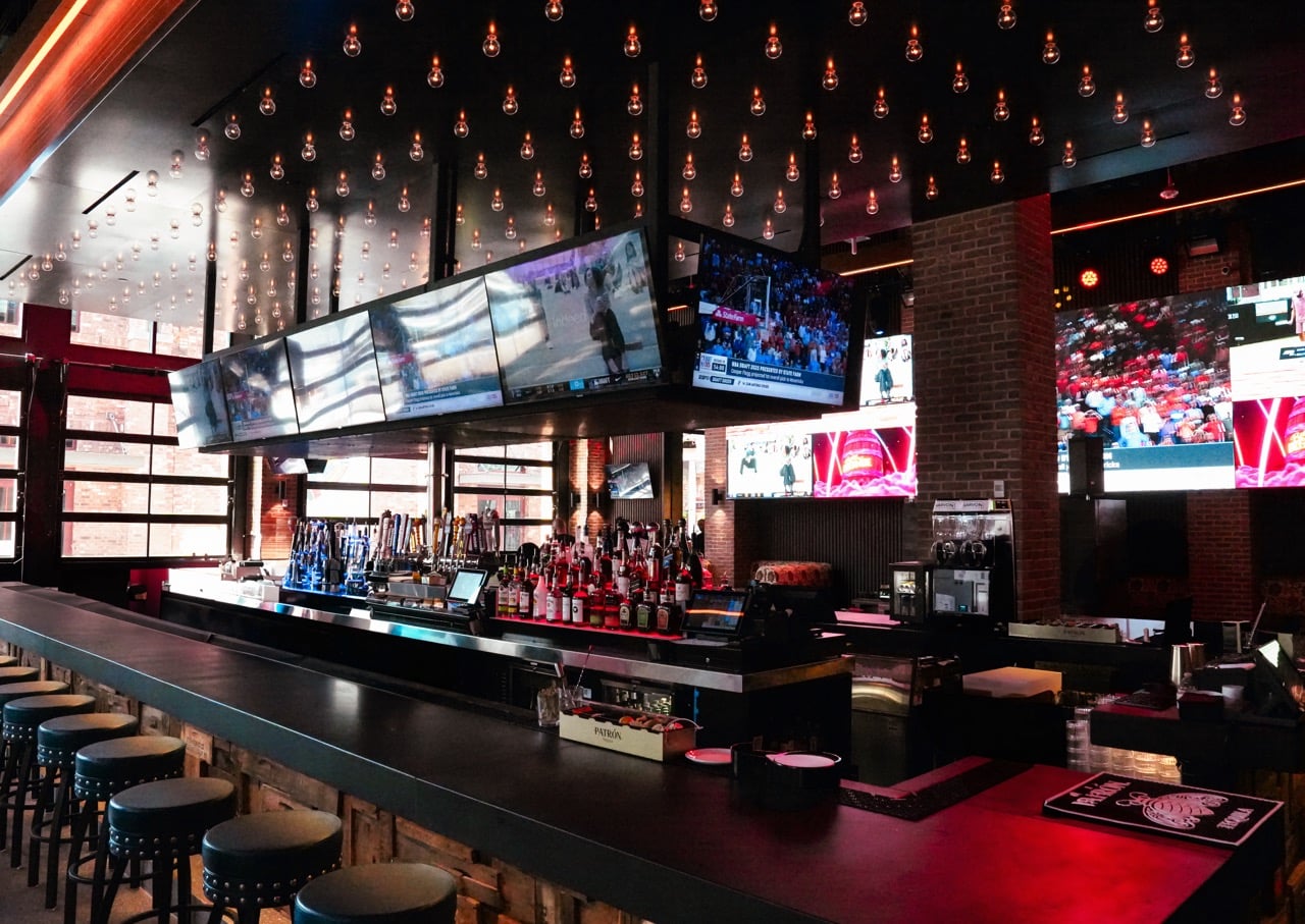 Long bar counter with liquor bottles multiple TV screens and hanging light bulbs