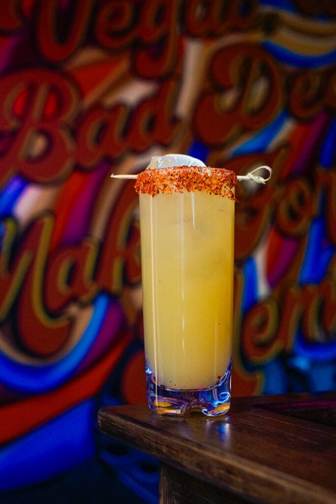 Tall yellow cocktail with tajin rim and ice garnished with a lime wedge