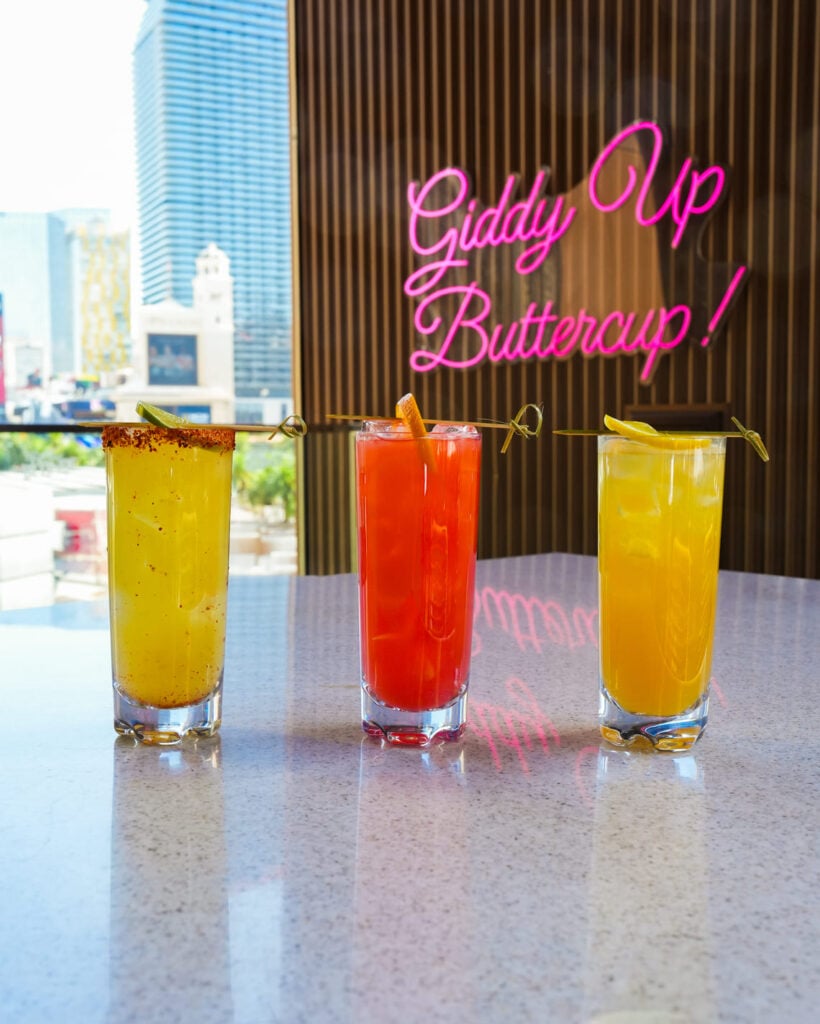 Three colorful cocktails on a marble counter with city view and neon sign