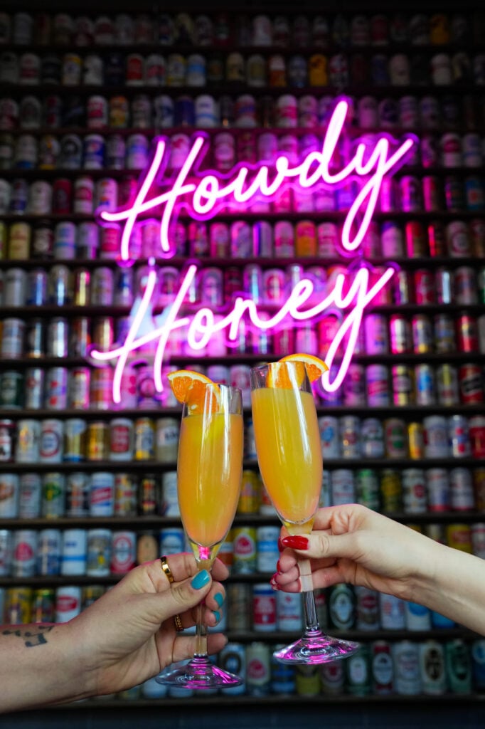 Two hands clinking mimosa glasses in front of neon Howdy Honey sign