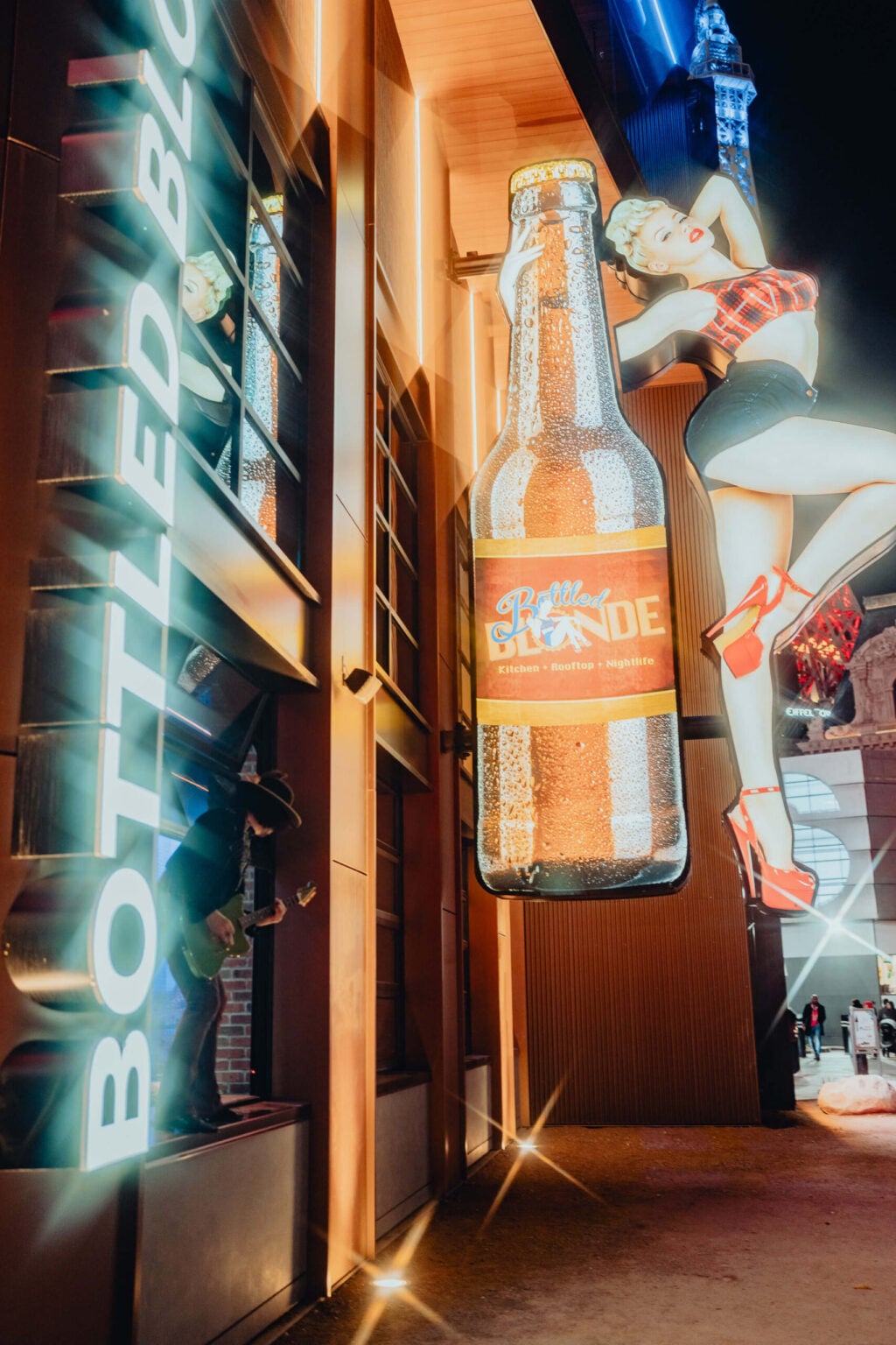 Exterior of Bottled Blonde Las Vegas at night featuring neon signage, a giant beer bottle display, and a retro pin-up style figure above the entrance.