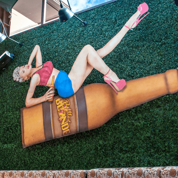 Pin-up style sculpture of a woman in a pink top, blue shorts, and pink high heels, lounging on a large "Bottled Blonde" beer bottle, mounted on a green artificial turf wall, with patterned outdoor seating below.