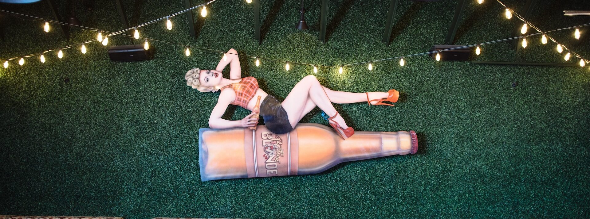 Pin-up style sculpture of a woman in a pink top, blue shorts, and pink high heels, lounging on a large "Bottled Blonde" beer bottle, mounted on a green artificial turf wall, with patterned outdoor seating below.
