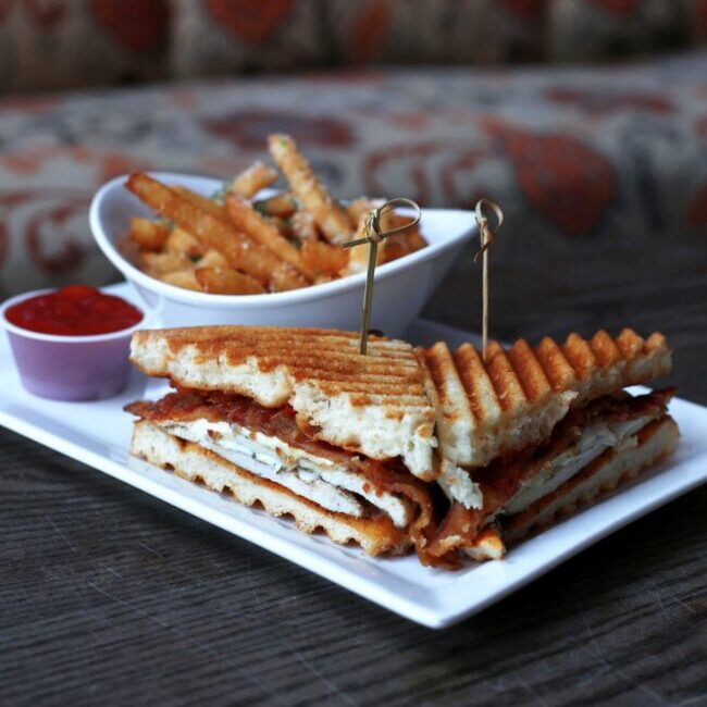 Grilled panini sandwich with crispy bacon and grilled chicken, served on a white plate with a side of golden French fries and a cup of marinara sauce.