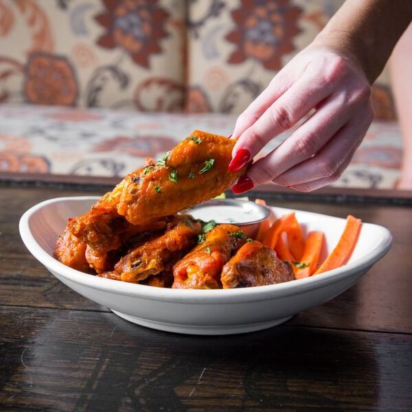 Person with red nails picking up a buffalo chicken wing from a white bowl filled with wings, carrot sticks, and a cup of ranch dressing.