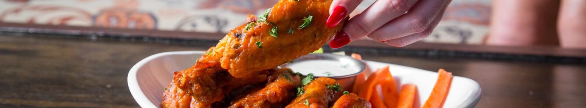 Person with red nails picking up a buffalo chicken wing from a white bowl filled with wings, carrot sticks, and a cup of ranch dressing.