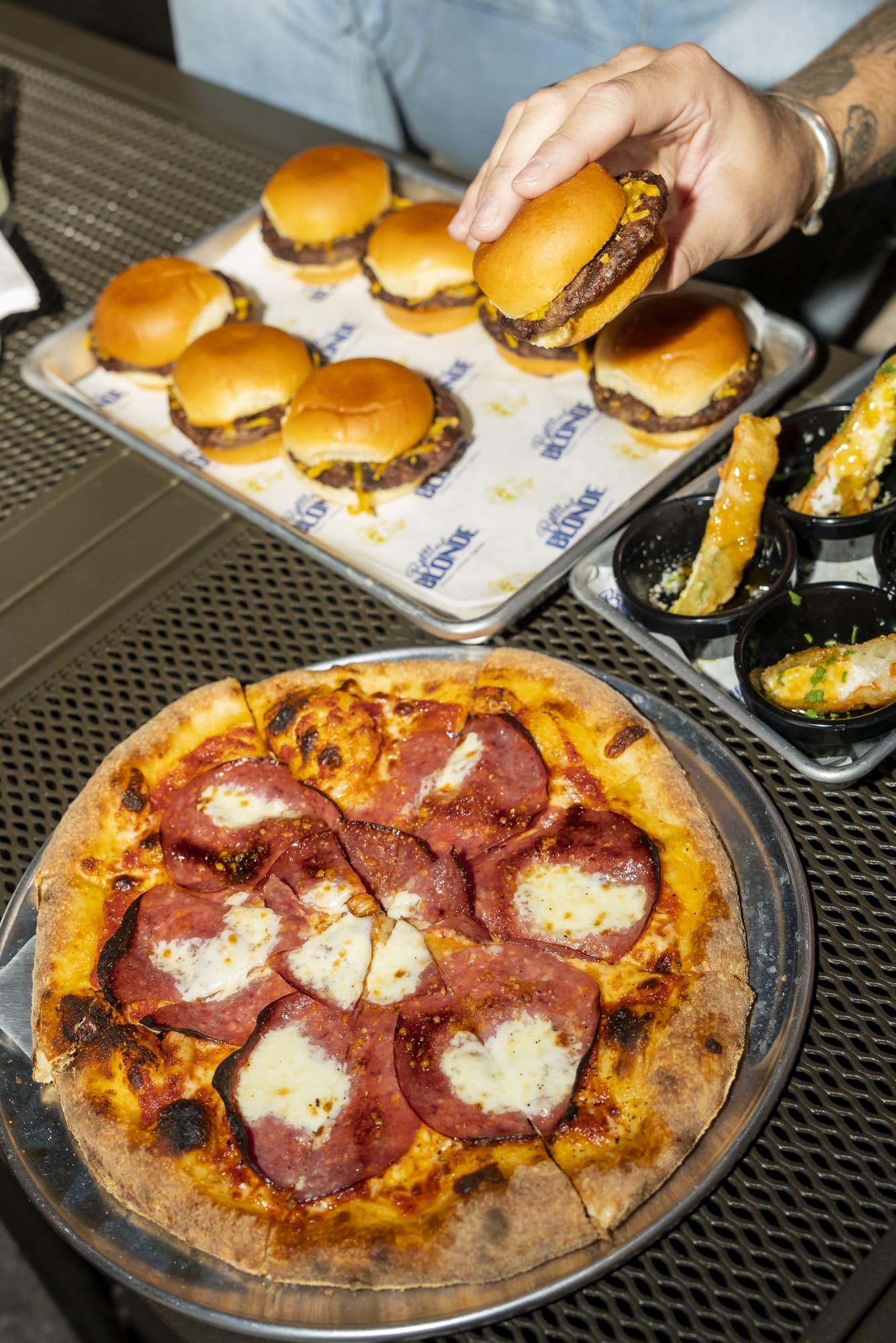 A pepperoni pizza sits on a metal tray beside mini cheeseburgers and fried appetizers at an outdoor dining table.
