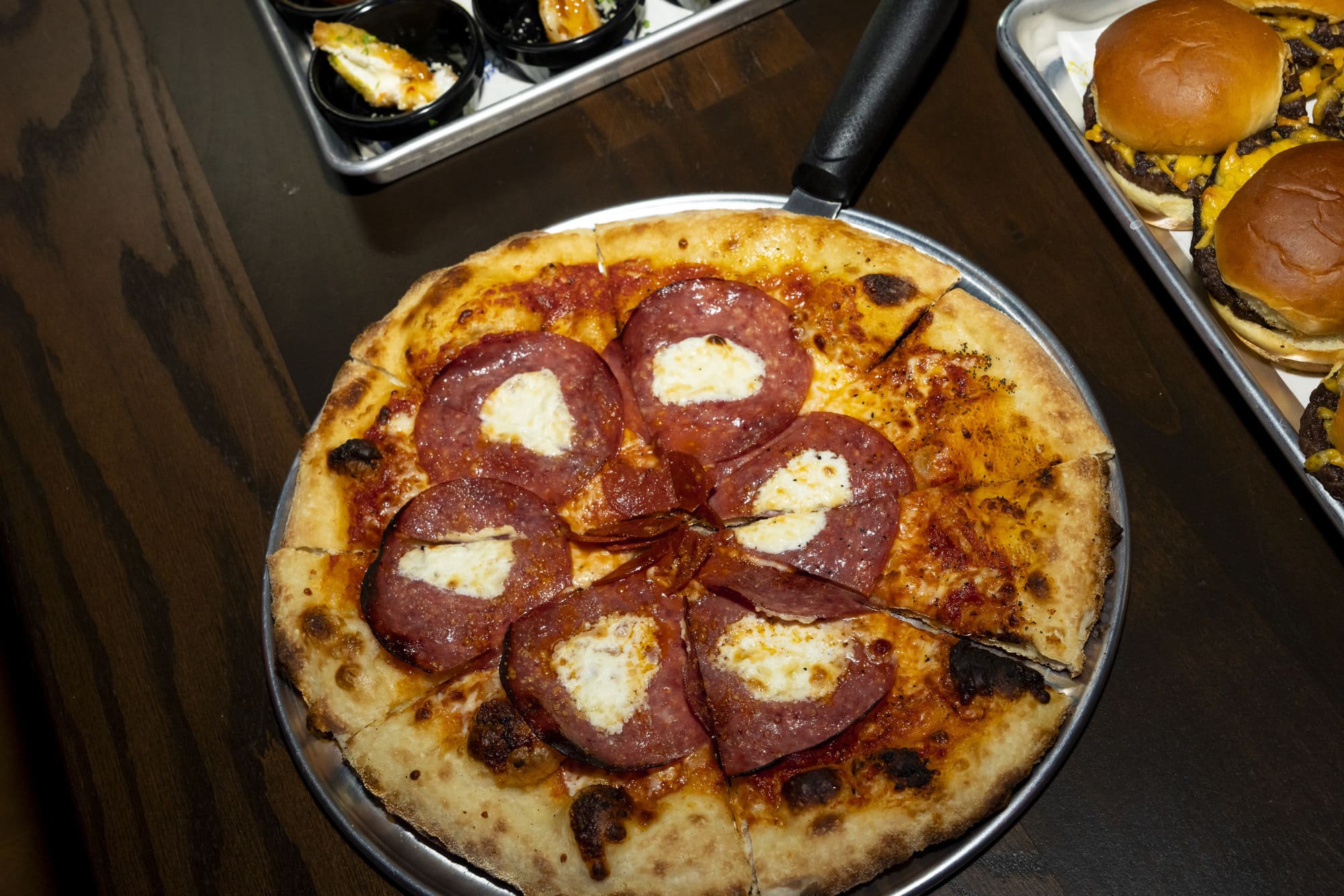 A pepperoni pizza and a tray of cheeseburgers on a dark wooden table at a restaurant.