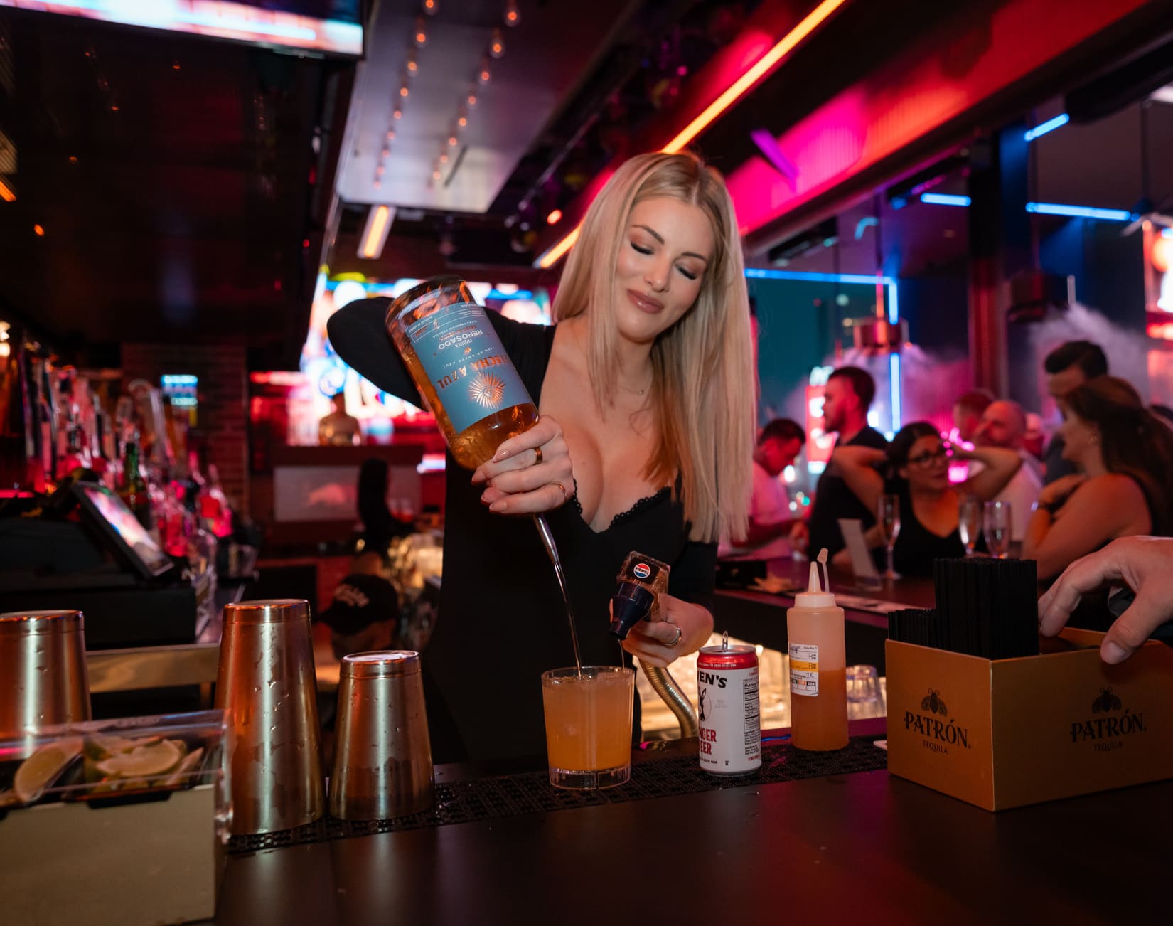 Female bartender pouring tequila into a cocktail at a busy nightclub bar.
