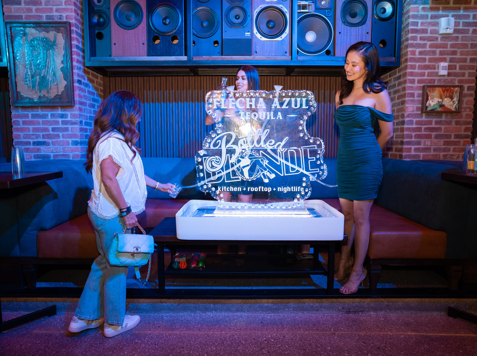 Three women gathered around a Bottled Blonde ice sculpture at a nightclub.