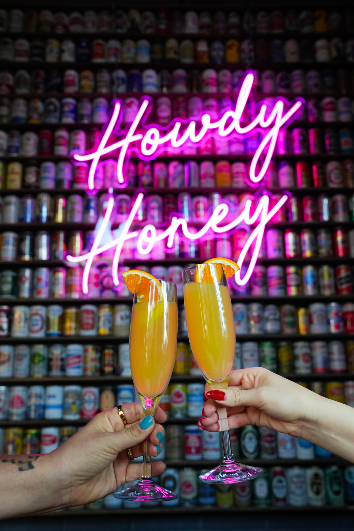 Two hands clinking mimosa glasses in front of neon Howdy Honey sign