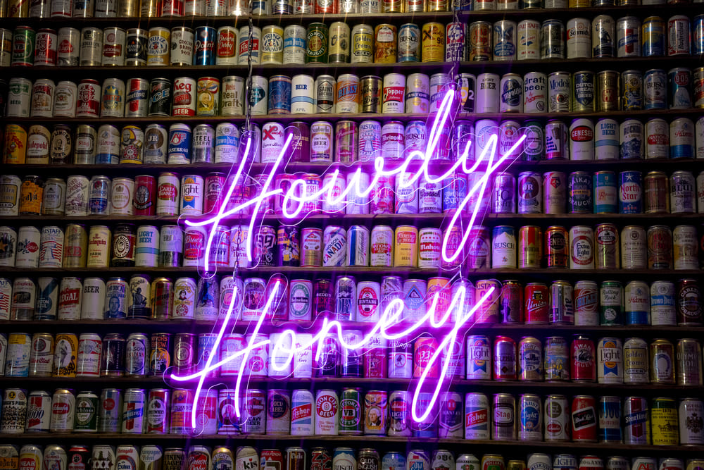Neon sign reading “Howdy Honey” in front of a wall of vintage beer cans