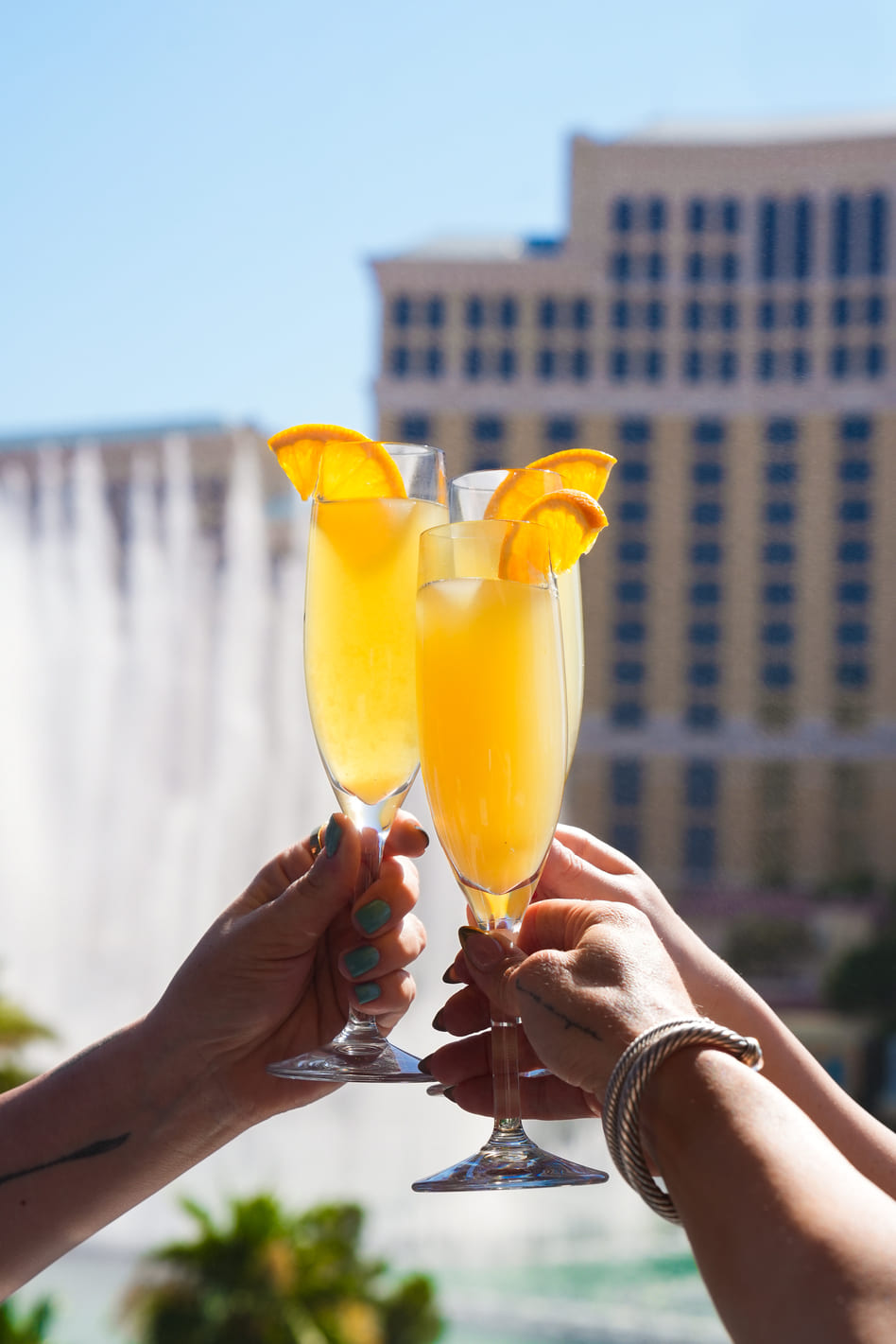 Three champagne flutes with orange slices held up in front of a hotel fountain