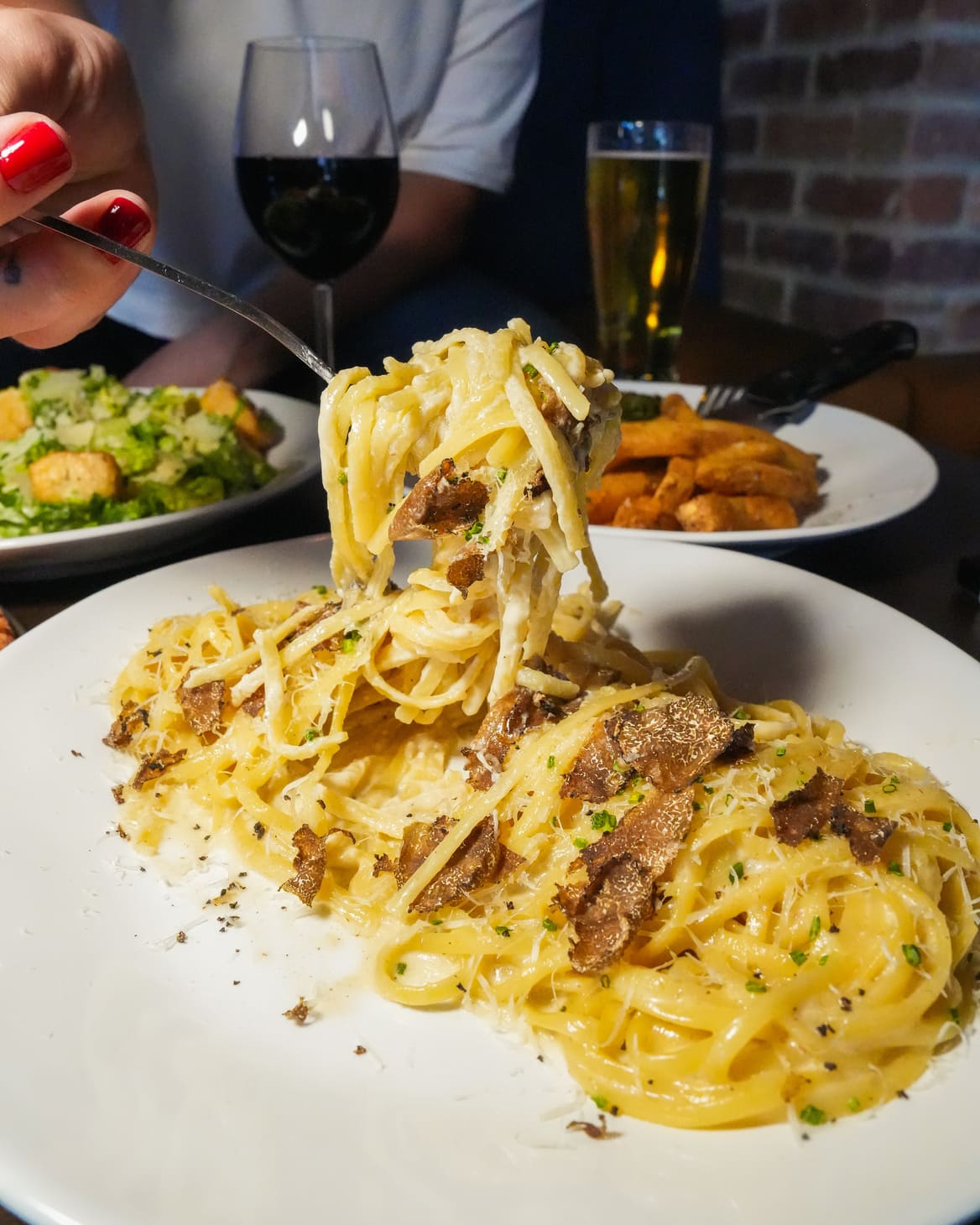 Fork lifting creamy truffle pasta with wine and fries in the background