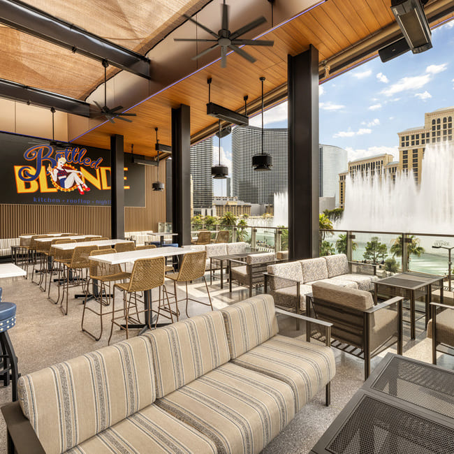Rooftop seating at Bottled Blonde Las Vegas with striped sofas, high-top tables, and a view of the Bellagio fountains.