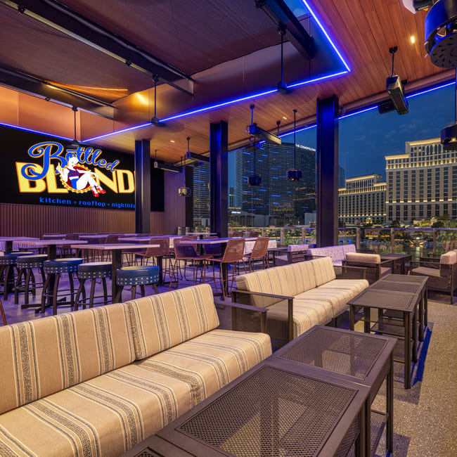 Rooftop lounge with striped cushioned seating, neon sign, and city skyline view at night.
