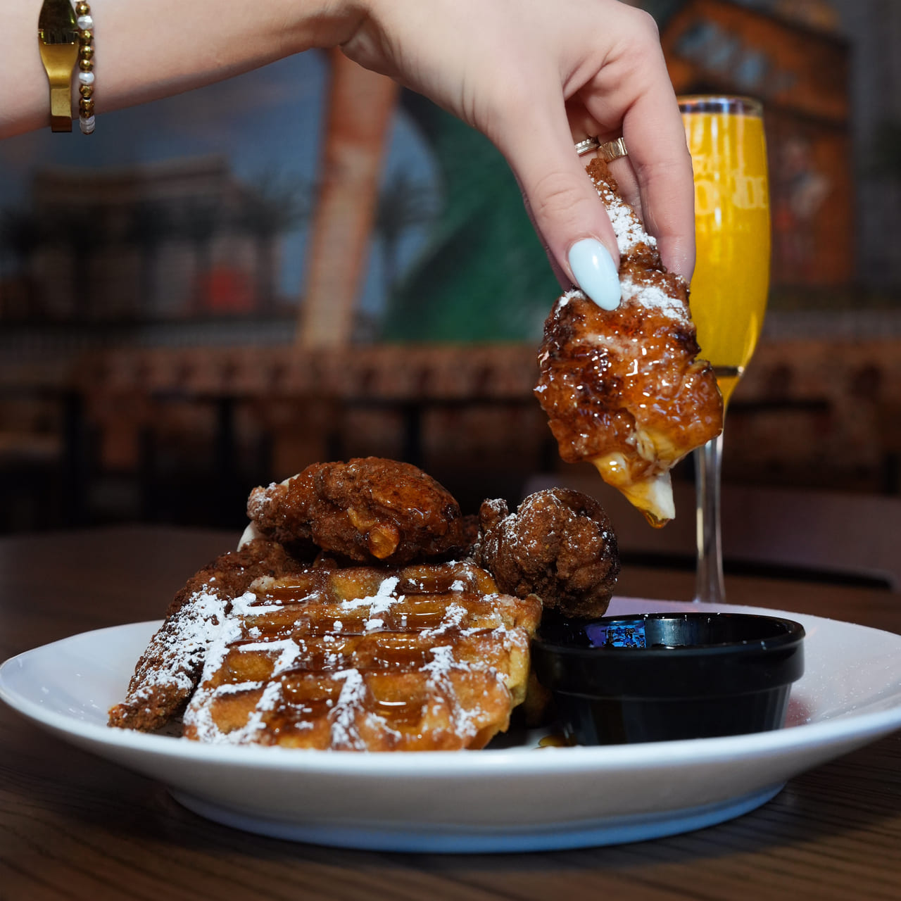 Hand dipping a piece of fried chicken into syrup, served with waffles dusted in powdered sugar and a mimosa in the background.