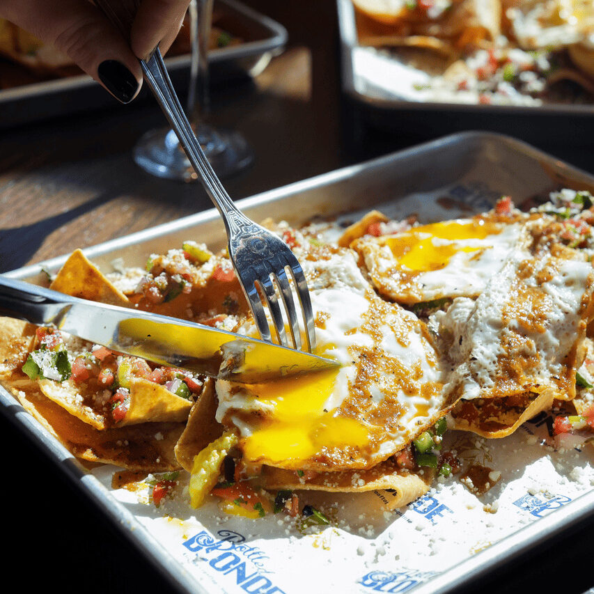 Close-up of chilaquiles topped with sunny-side-up eggs being cut with a fork and knife. A mimosa sits in the background on a sunlit table.