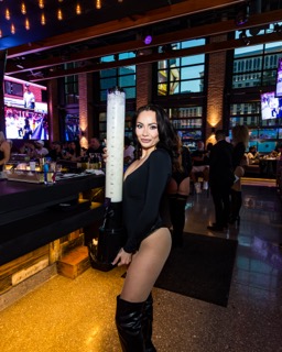 Woman in black outfit holding a tall beer tower inside a vibrant bar with TVs and colorful lighting.
