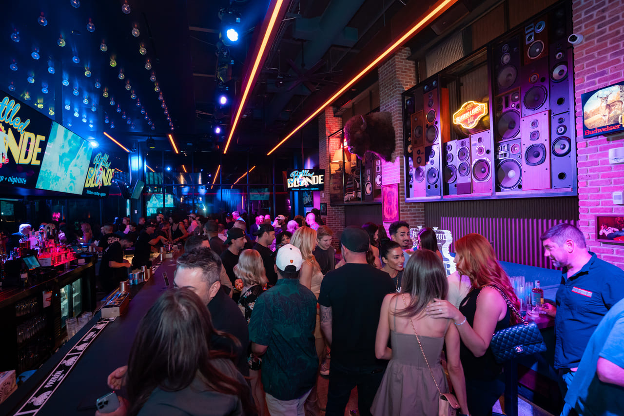 Crowded nightlife scene at Bottled Blonde with patrons socializing at the bar under neon and LED lights, surrounded by speakers and vibrant decor.