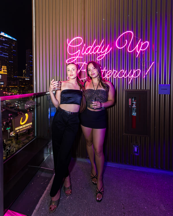Two women posing with drinks at night beside a neon sign reading "Giddy Up Buttercup!" at Bottled Blonde Las Vegas.