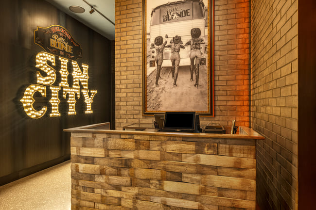 Reception area at Bottled Blonde Las Vegas with “Sin City” marquee lights and vintage photo on brick wall.