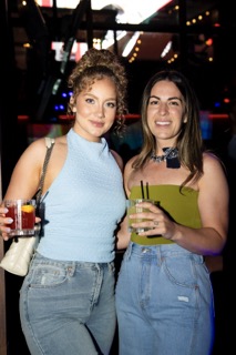 Two women smiling and holding cocktails at a nightlife venue with colorful lighting in the background.