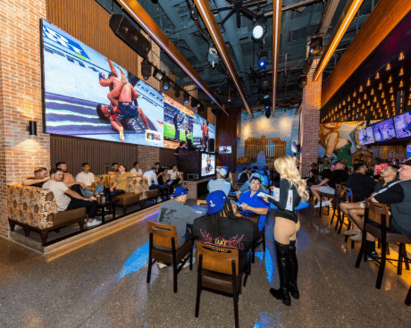 Guests watching a UFC match on large wall-mounted screens inside a lively sports bar with a waitress serving drinks.