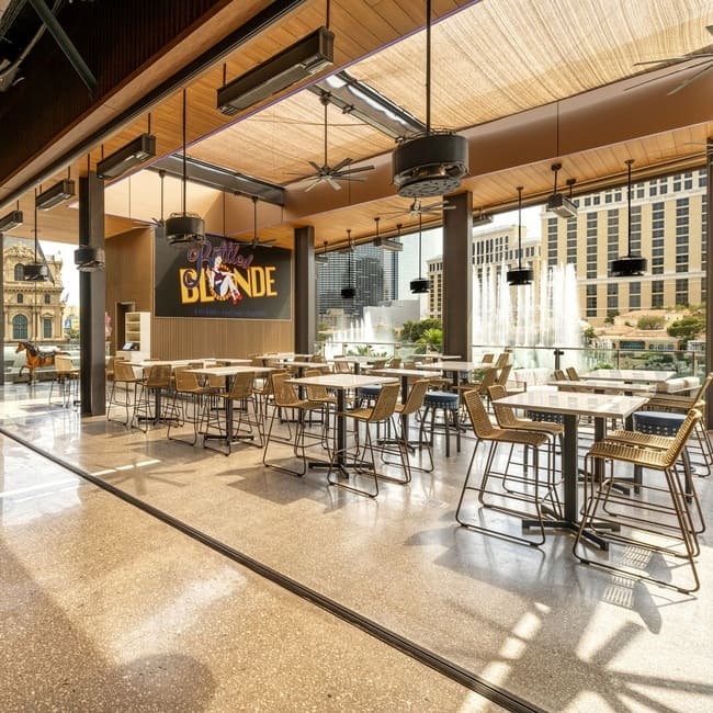 Indoor seating at Bottled Blonde Las Vegas with high-top tables, wicker chairs, and Bellagio fountain views.