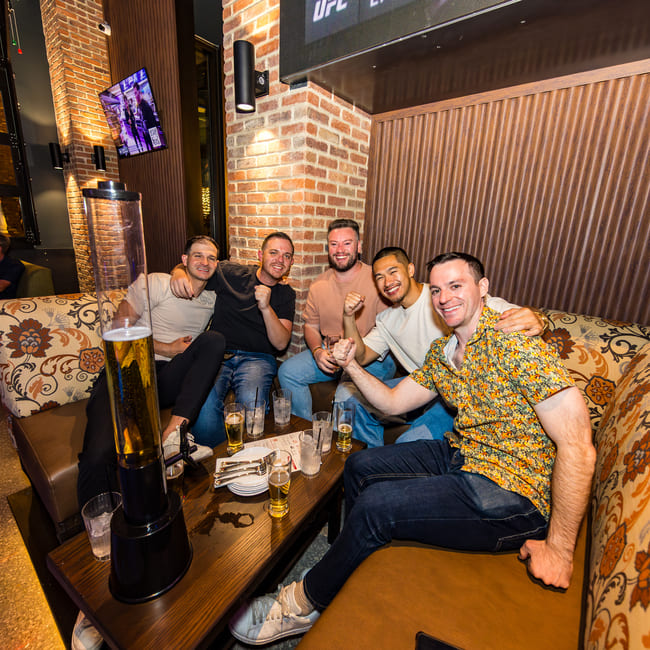 Group of five men sitting in a booth with drinks, smiling and posing with fists up inside a sports bar.