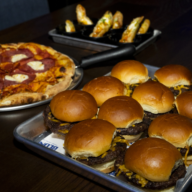 Tray of cheeseburger sliders with a pepperoni pizza and stuffed breadsticks on a dark wooden table.