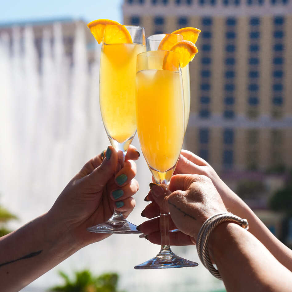Three champagne flutes with orange slices held up in front of a hotel fountain