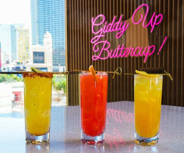 Three colorful cocktails on a marble counter with city view and neon sign