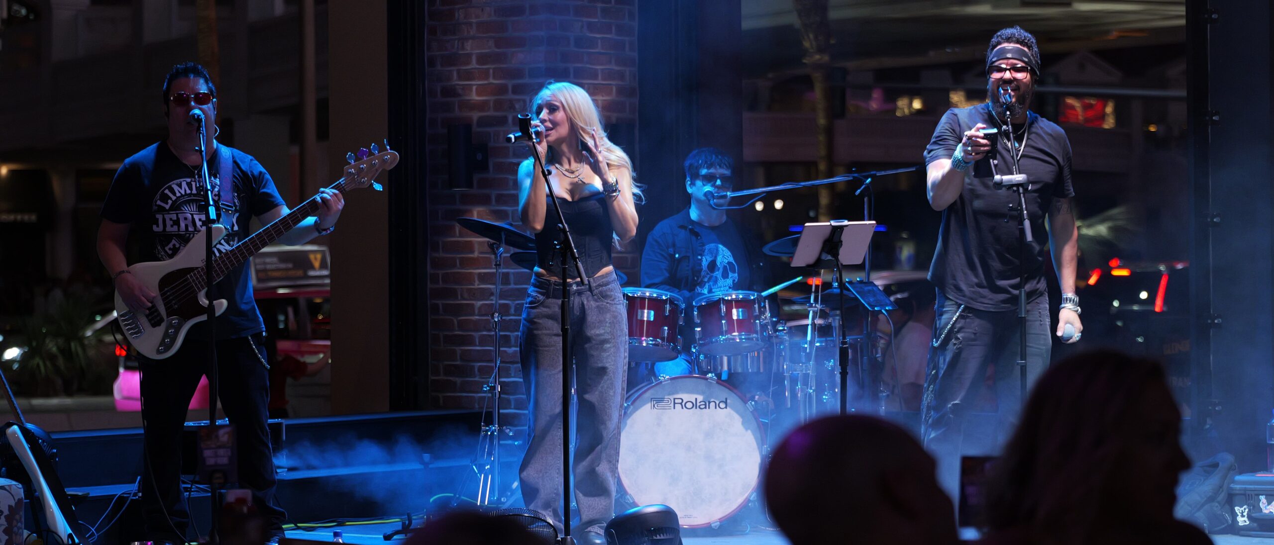 Live band performing at night with male and female vocalists on stage, drummer and guitarist in background, and smoke effects enhancing the atmosphere.
