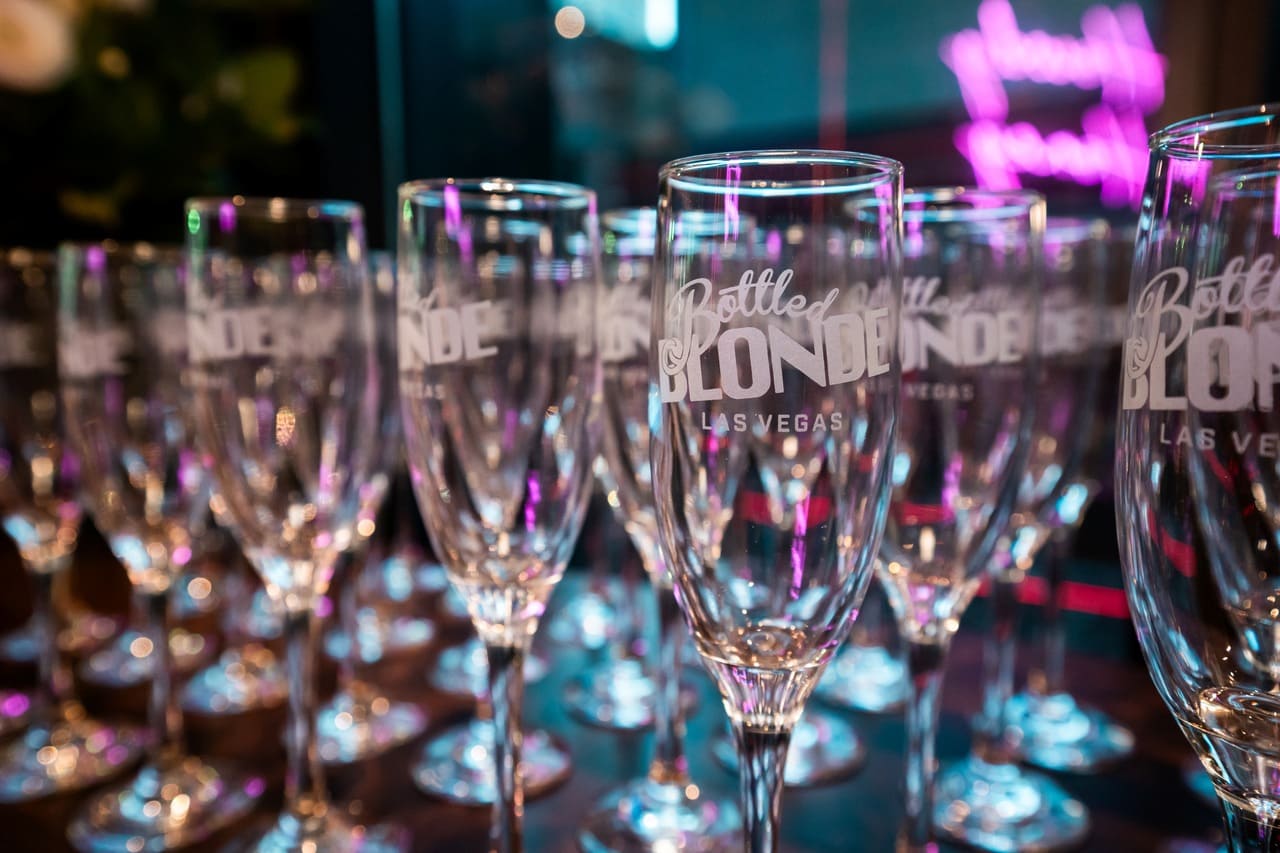 Close-up of champagne flutes etched with the Bottled Blonde Las Vegas logo, arranged in rows with colorful lights reflecting on the glass.