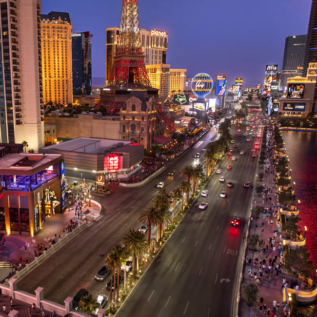 Aerial view of the Las Vegas Strip at dusk, featuring the Paris Las Vegas hotel with its illuminated Eiffel Tower replica and busy traffic lined with palm trees.