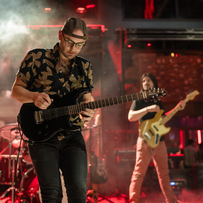 Male guitarist performing live on stage in a dimly lit venue with red lighting, wearing a printed shirt and backwards cap, while a bassist plays in the background.