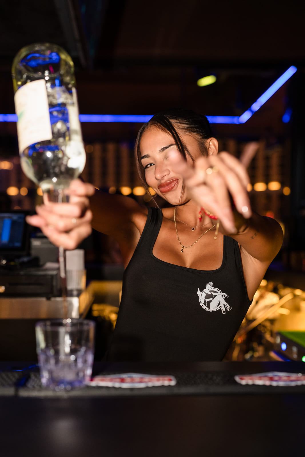 Smiling female bartender holding up a bottle while pouring a drink at a neon-lit bar.