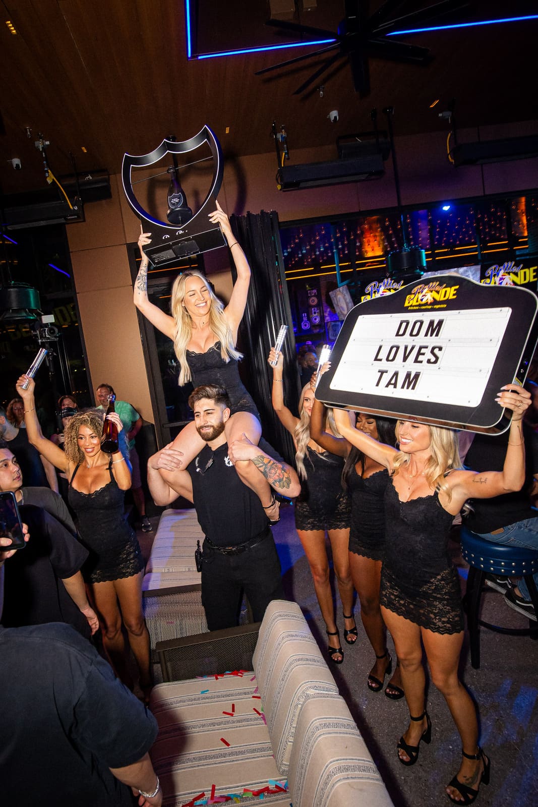 Woman on shoulders holding bottle display as group holds light-up signs.