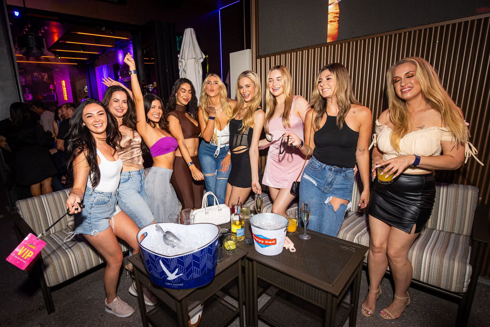 Group of women in casual nightlife attire smiling and posing on a rooftop patio with drink buckets.