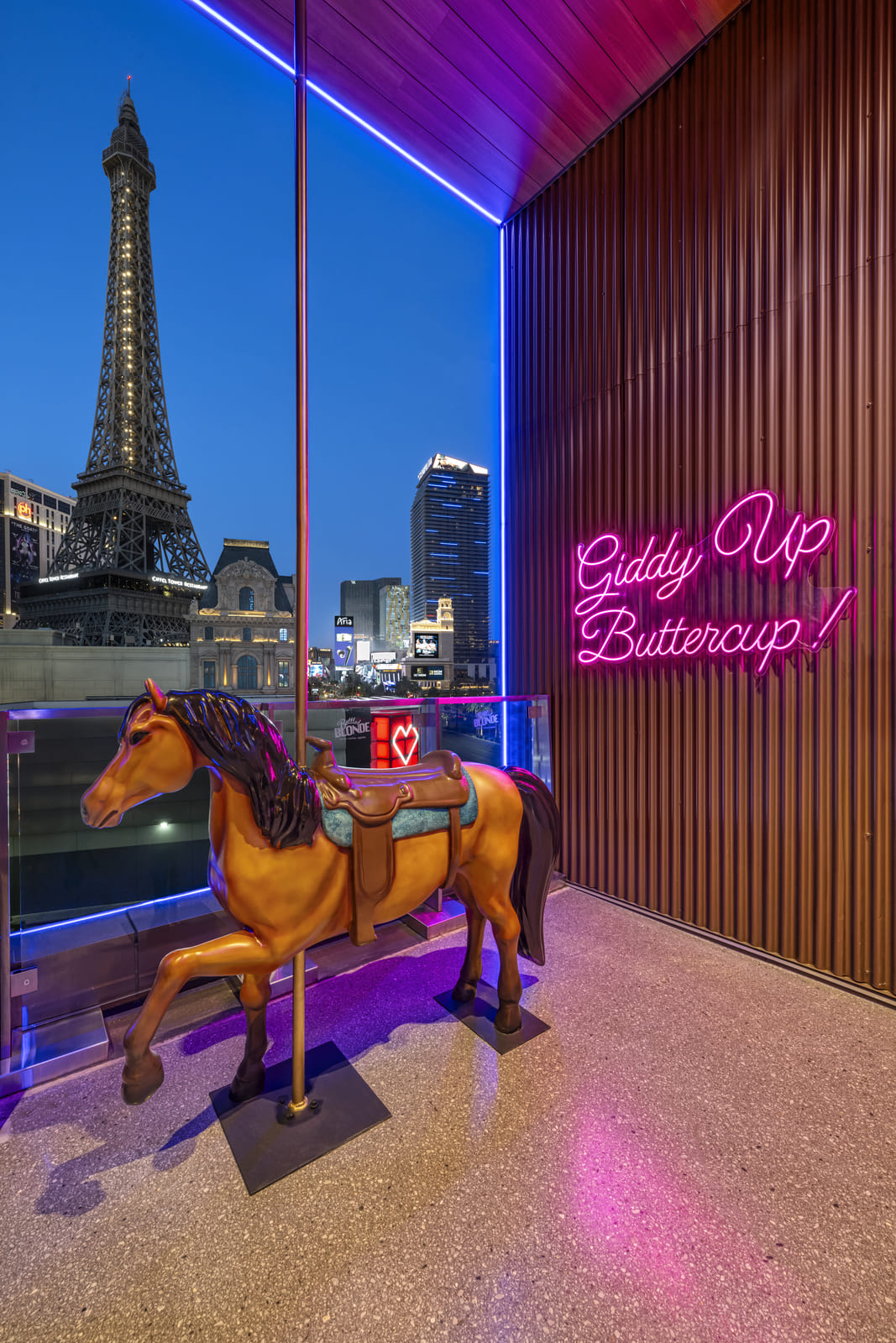 Life-sized mechanical horse next to neon sign that says “Giddy Up Buttercup!” with Eiffel Tower in background.