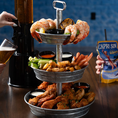 Three-tier tray with shrimp, chicken wings, fries, and sauces, served with beer and a Capri Sun cocktail pouch.