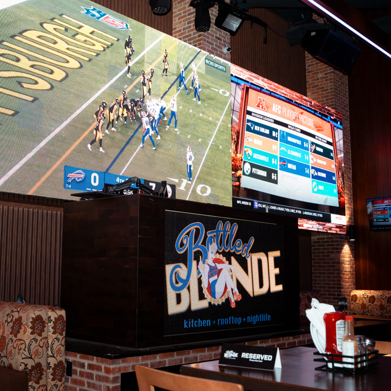 Large screen shows a live football game between the Steelers and Bills inside Bottled Blonde bar.