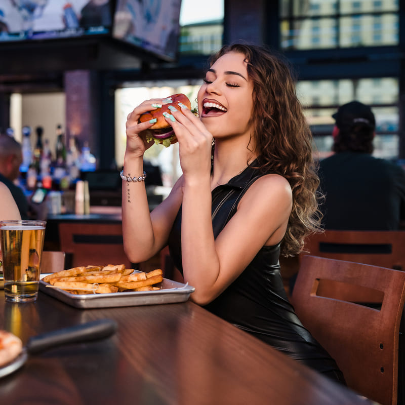 Smiling woman in a black outfit about to take a bite of a burger, with fries and beer in front of her at Bottled Blonde.